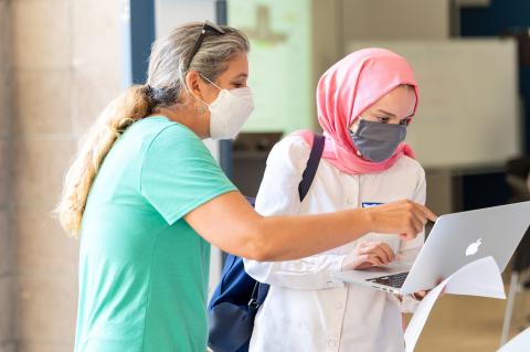 two masked people standing and working on a Mac laptop