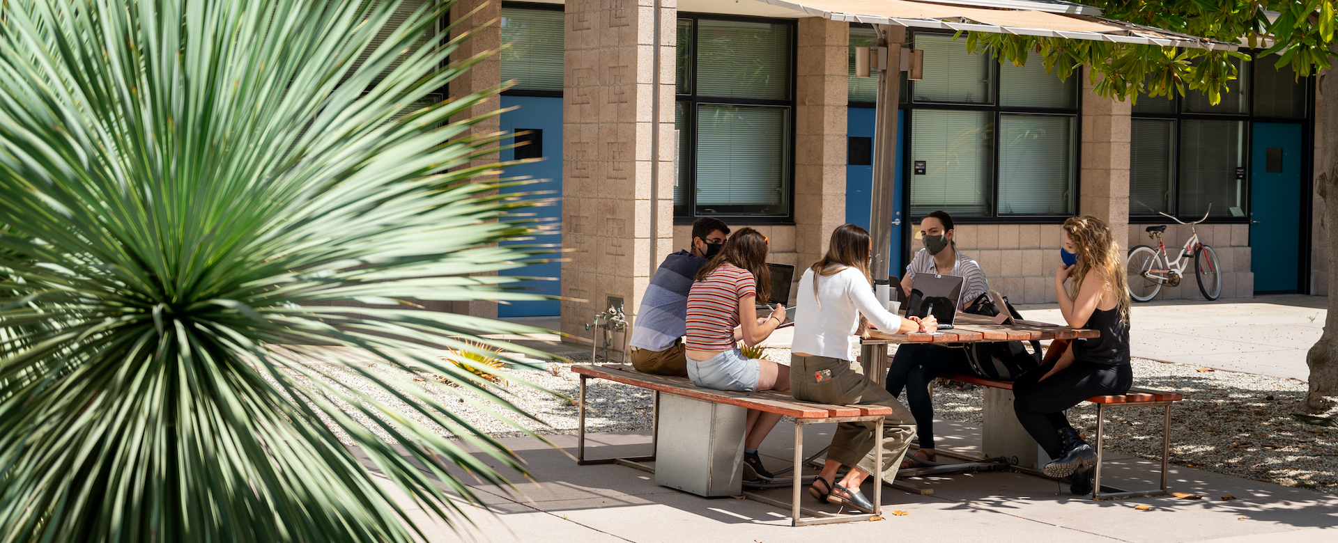 photo of five people masked and seated at a picnic bench in a courtyard