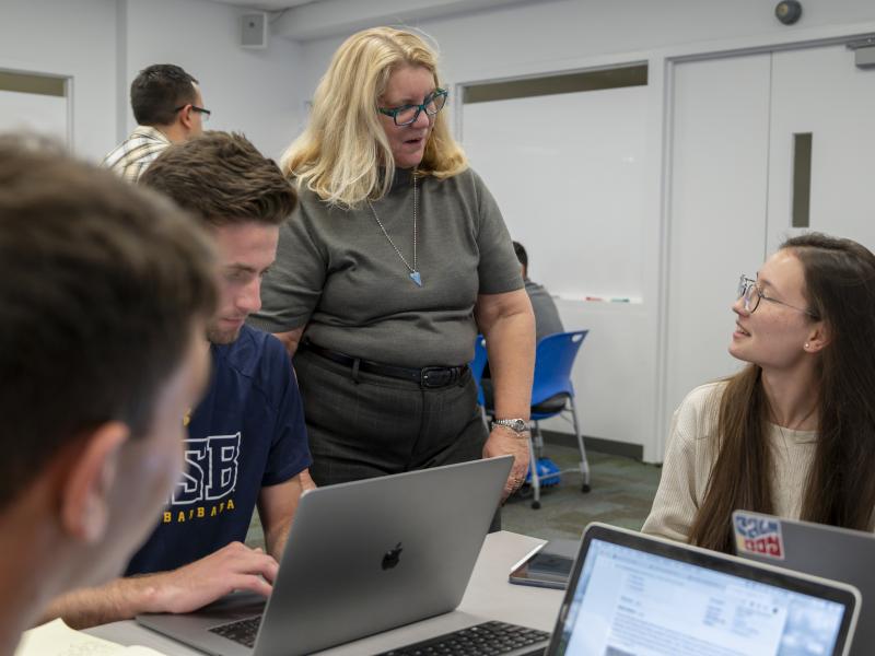 blonde person standing next to student sitting at a desk both conversing