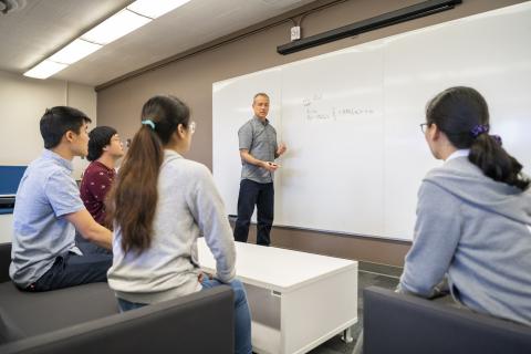 photo of standing person at a whiteboard talking to four seated people