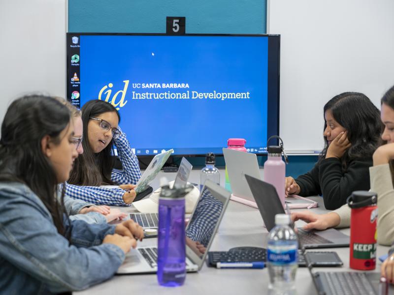 photo with four visible students working on laptops around a table in front of a tv screen