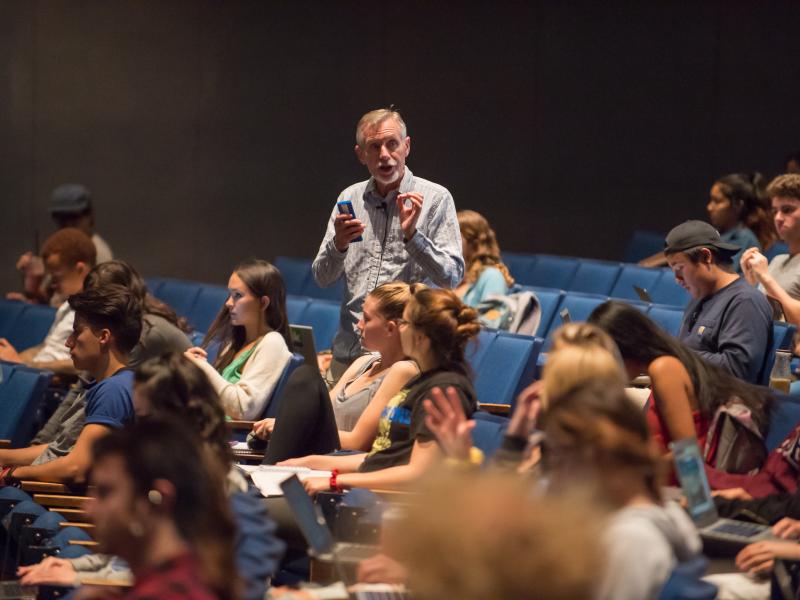 photo with a side view of students in a lecture hall with a teacher standing in the aisle